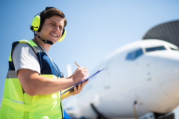 Looking positive. Aviation technician looking at camera with smile and writing information on clipboard. Passenger airplane on blurred background