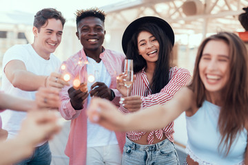 Portrait of laughing girls and cheerful men tasting appetizing alcohol liquid while holding bengal lights in arms