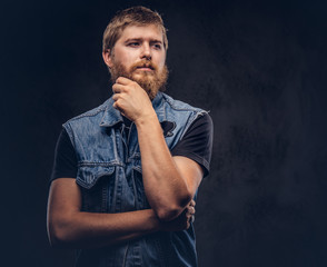 Portrait of a pensive hipster guy dressed in jeans jacket posing with hand on chin on a dark background.