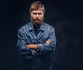 Portrait of a handsome hipster guy dressed in jeans jacket posing with crossed arms on a dark background.