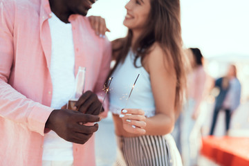 Smiling lady hugging glad man. He tasting bottle of liquid. They holding bengal lights in hands