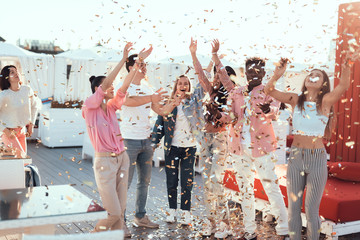 Satisfied ladies and beaming males catching confetti while having fun during funny party outdoor - obrazy, fototapety, plakaty