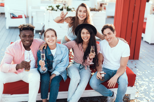 Portrait of beaming girls and cheerful males looking at camera while tasting glasses of champagne and bottles of beer. Glad lady wearing modern jacket. Positive friends during rest concept