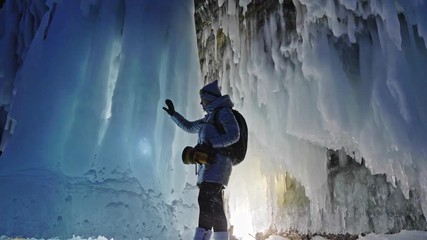 Travel woman on ice cave of Lake Baikal. Trip to winter island. Girl backpacker is walking of ice grot. Traveler looks at beautiful ice grotto. Hiker wears sports glasses, silver jacket, backpack, led - Powered by Adobe