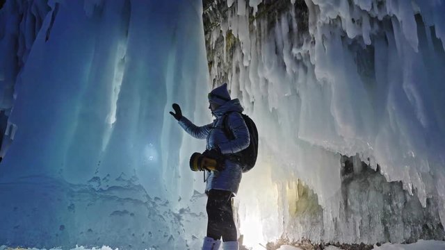 Travel woman on ice cave of Lake Baikal. Trip to winter island. Girl backpacker is walking of ice grot. Traveler looks at beautiful ice grotto. Hiker wears sports glasses, silver jacket, backpack, led
