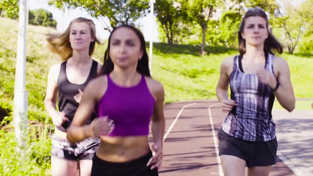 Dolly shot of three young women running along the path for runners at the sports ground. Front view