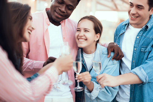 Smiling man embracing beaming lady while telling with happy friends. They drinking glasses of champagne during party