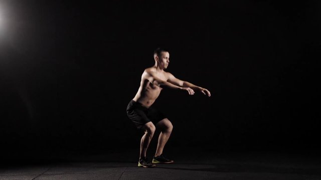 young strong man with naked torso is doing squatting in a fitness hall in evening, stretching hands forward