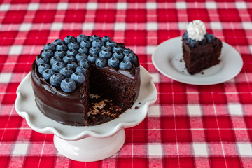 Round chocolate cake topped with fresh blueberries, on a white ceramic cake plate, red and white plaid tablecloth, slice on white dessert plate with whipped cream