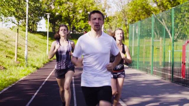 Dolly shot of three young people running along the path for runners at the sports ground. Front view
