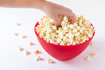 Fototapeta na wymiar Hand reaching for popped popcorn in a big red plastic bowl on a white table, with spilled kernels