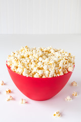 Vertical of popped popcorn in a big red plastic bowl on a white table, with spilled kernels