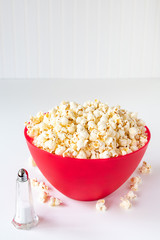 Vertical of popped popcorn in a big red plastic bowl on a white table, with a glass salt shaker and spilled kernels