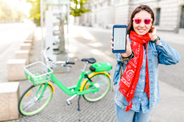 Attractive happy woman using smart phone and rent mobile application to share modern green electric bicycle, alternative eco transport concept