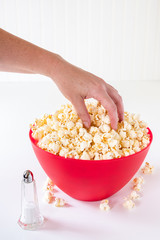 Vertical of hand reaching for popped popcorn in a big red plastic bowl on a white table, with a glass salt shaker and spilled kernels