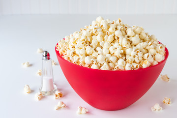 Popped popcorn in a big red plastic bowl on a white table, with a glass salt shaker and spilled kernels