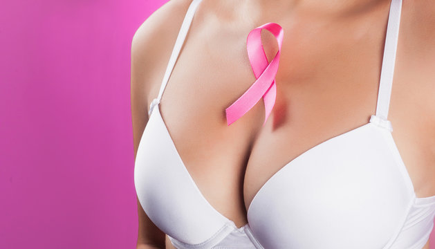 Beautiful female breast in a white bra with a pink ribbon on her chest. The concept of support for cancer patients. Close-up - pink background