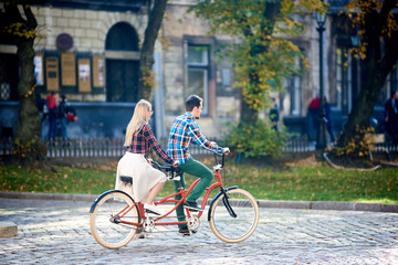 Young active couple, bearded man and long-haired blond woman riding tandem bicycle along paved city street on bright sunny summer or early autumn day on blurred trees and buildings background.