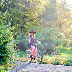 Slim blond fashionable long-haired attractive girl in glasses, short dress and pink hat riding lady bicycle along paved park alley on beautiful green and golden trees lit by bright sun background.