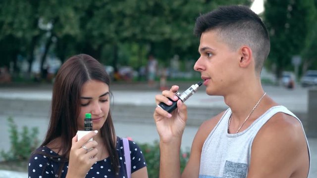 Portrait of a young couple from a smiling girl in a polka-dot dress and a young man in a singlet smoking e-cigarettes and chatting in summer