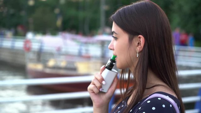 Profile of a thoughtful girl standing and smoking an e-cigarette on a river quay with yachts at splendid sunset. She is in dense smoke