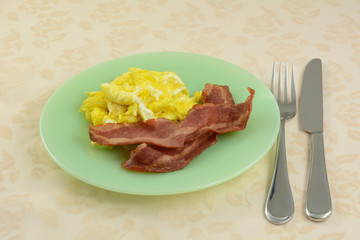 Quick breakfast of scrambled eggs and two strips of turkey bacon on green plate with fork and knife