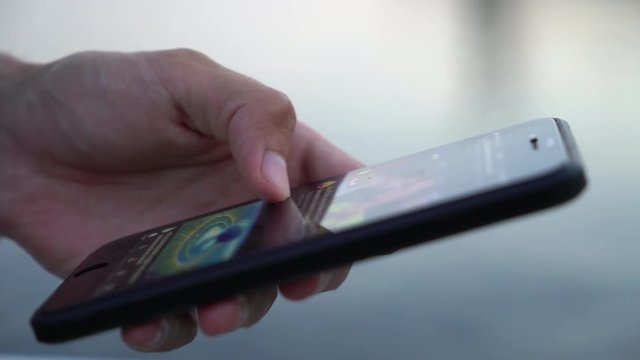 Closeup of a man`s hand keeping a black phone and touching its screen while scrolling and seeking some information on a river quay in summer