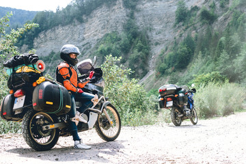 A girl dressed in body armor and helmet stand alone with big adventure touring motorcycle with bags and camping equipment, off road travel jorney, traveling together, couple. mountain dirt road