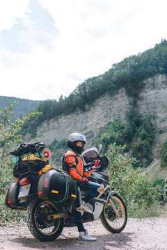 A girl dressed in body armor and helmet stand alone with big adventure touring motorcycle with bags and camping equipment, off road travel jorney, traveling together. mountain dirt road vertical photo
