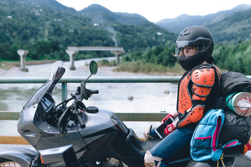 Fototapeta na wymiar A girl in body armor and helmet sit on a big adventure touring motorcycle with bags and camping equipment, off road travel jorney, traveling together, couple, bridge, river mountains background