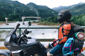 Fototapeta na wymiar A girl in body armor and helmet sit on a big adventure touring motorcycle with bags and camping equipment, off road travel jorney, traveling together, couple, bridge, river mountains background