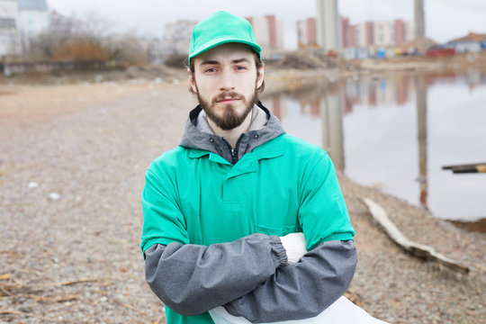 Young bearded volunteer in green uniform standing with arms crossed and looking at camera on shore