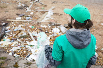 Back view of volunteer man in green uniform standing with bag at pile with trubbish