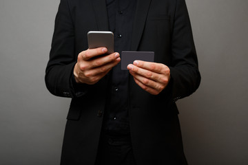 Young businessman holding a phone and a credit card.Man dressed in black suit on gray background