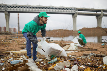 Young bearded man in green uniform collecting litter on coast