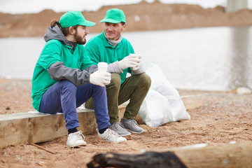 Two volunteers in green uniform sitting with paper cups and resting on polluted shore