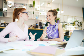 Cheerful pretty brainstorming women sitting at desk with laptop and papers