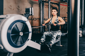 Naklejka na ściany i meble Fit woman working out on row machine in gym. Workout woman cross training exercising cardio using rowing machine in fitness gym.