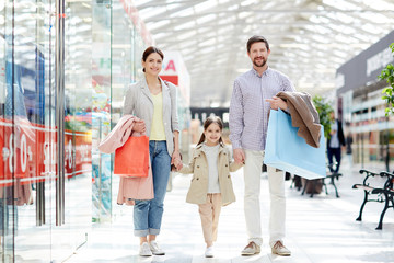 Obraz premium Adult cheerful parents with little girl carrying paper bags and standing in mall smiling at camera enjoying shopping together