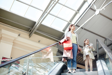 From below shot of adult woman with little girl standing on moving stairs in shopping mall carrying paper bags