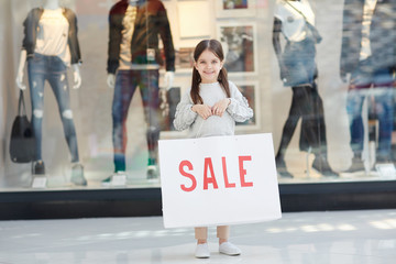 Adorable girl with ponytails smiling and looking at camera while holding banner with sale writing...