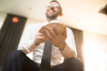 From below shot of adult smiling businessman holding white cup of coffee on saucer sitting in leisure