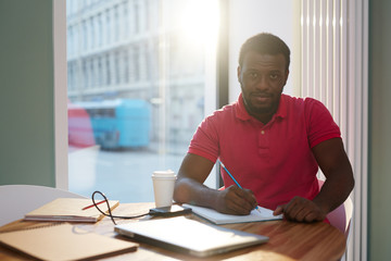 Casual black man sitting with coffee and papers at table taking notes in notepad and looking at camera in back lit