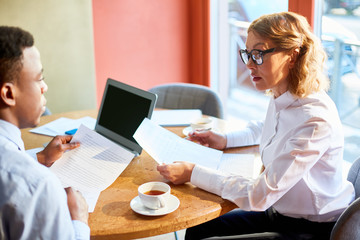 Diverse serious man and woman having business meeting and working with papers while sitting at table with tea cups