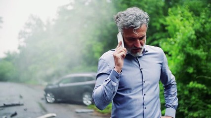 Mature man making a phone call after a car accident, smoke in the background.