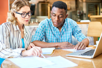 Obraz premium Attractive adult woman pointing at documents for black man while sitting at cafe table and working on project together