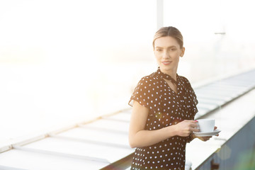 Elegant woman in polka dotted blouse holding cup of coffee and looking at camera in bright light standing outdoors