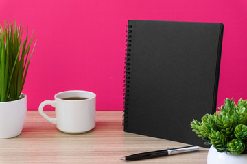 Black notebook with coffee cup and potted plant on wood desk with pink background. Text copy space.