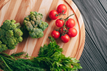 On a dark wooden table, on a round wooden board fresh green broccoli, parsley, dill and cherry tomatoes for your health. Recipe. Ingredients. Dietary food. Place under the text. View from above.