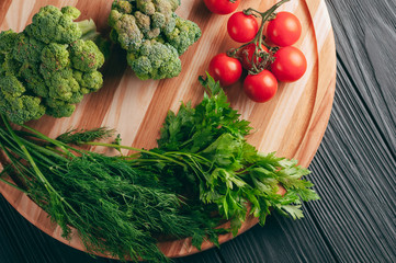 On a dark wooden table, on a round wooden board fresh green broccoli, parsley, dill and cherry tomatoes for your health. Recipe. Ingredients. Dietary food. Place under the text. View from above.